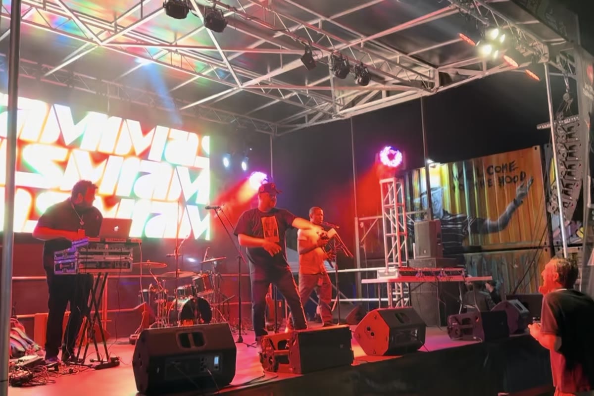 A band performing on an outdoor stage at night, with a lit LED screen beside them under a truss roof.