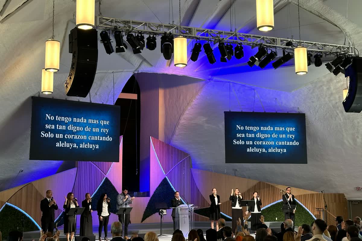 Two LED screens hung either side of a stage in a high white-roofed venue, both showing the same Spanish lyric slide, with a group singing below.