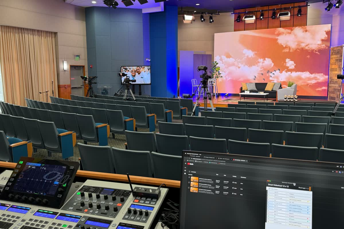 An LED wall behind a stage set with a sofa and side table, seen from a production position with an audio console and broadcast cameras in the foreground.