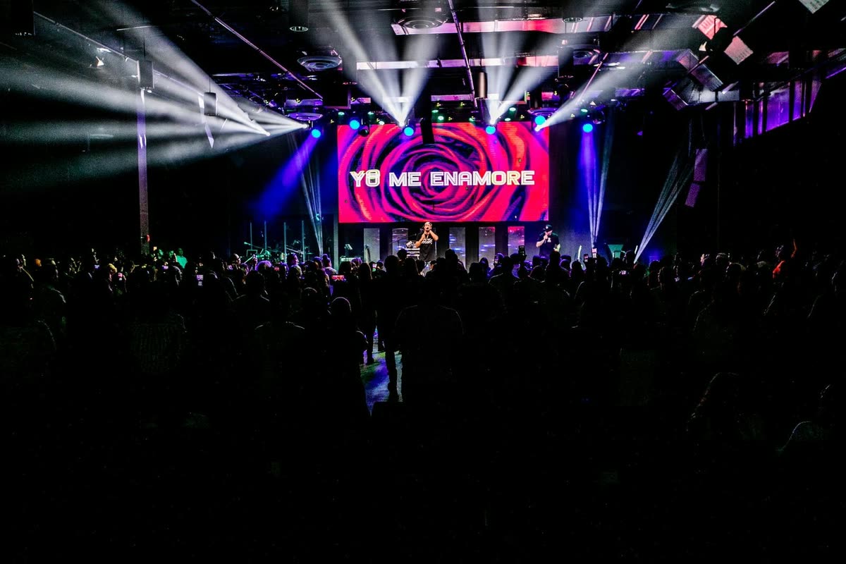 A packed dark venue facing a lit LED screen showing a magenta rose graphic and the words Yo Me Enamore, with beams of light over the crowd.