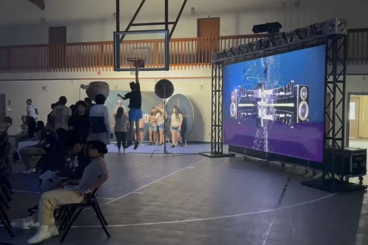 An LED wall in a ground-supported truss frame seen from across a gymnasium, with an audience seated on folding chairs in front of it.