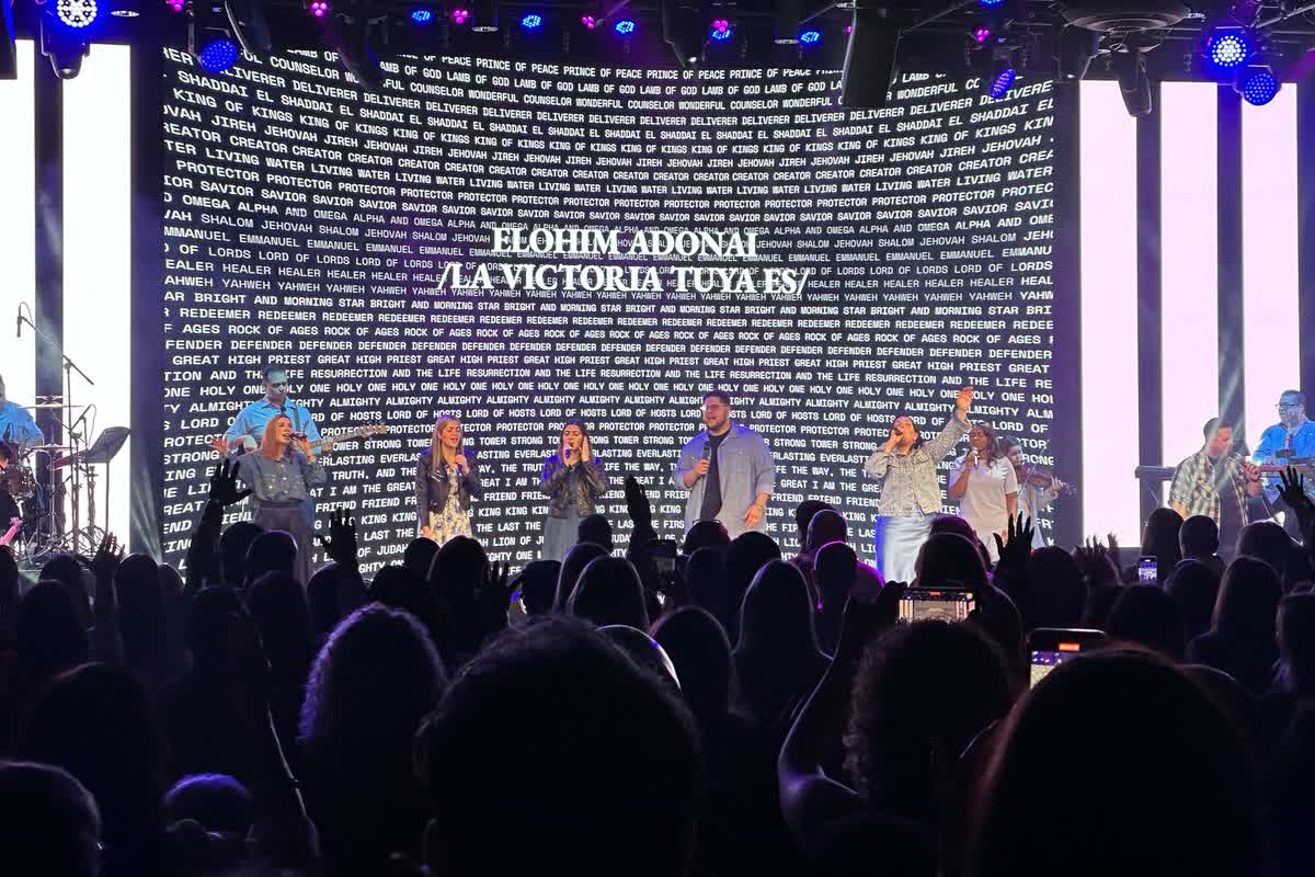 A close view from the audience of an LED wall filling the frame with white lettering over a blue background, singers on the stage below it.