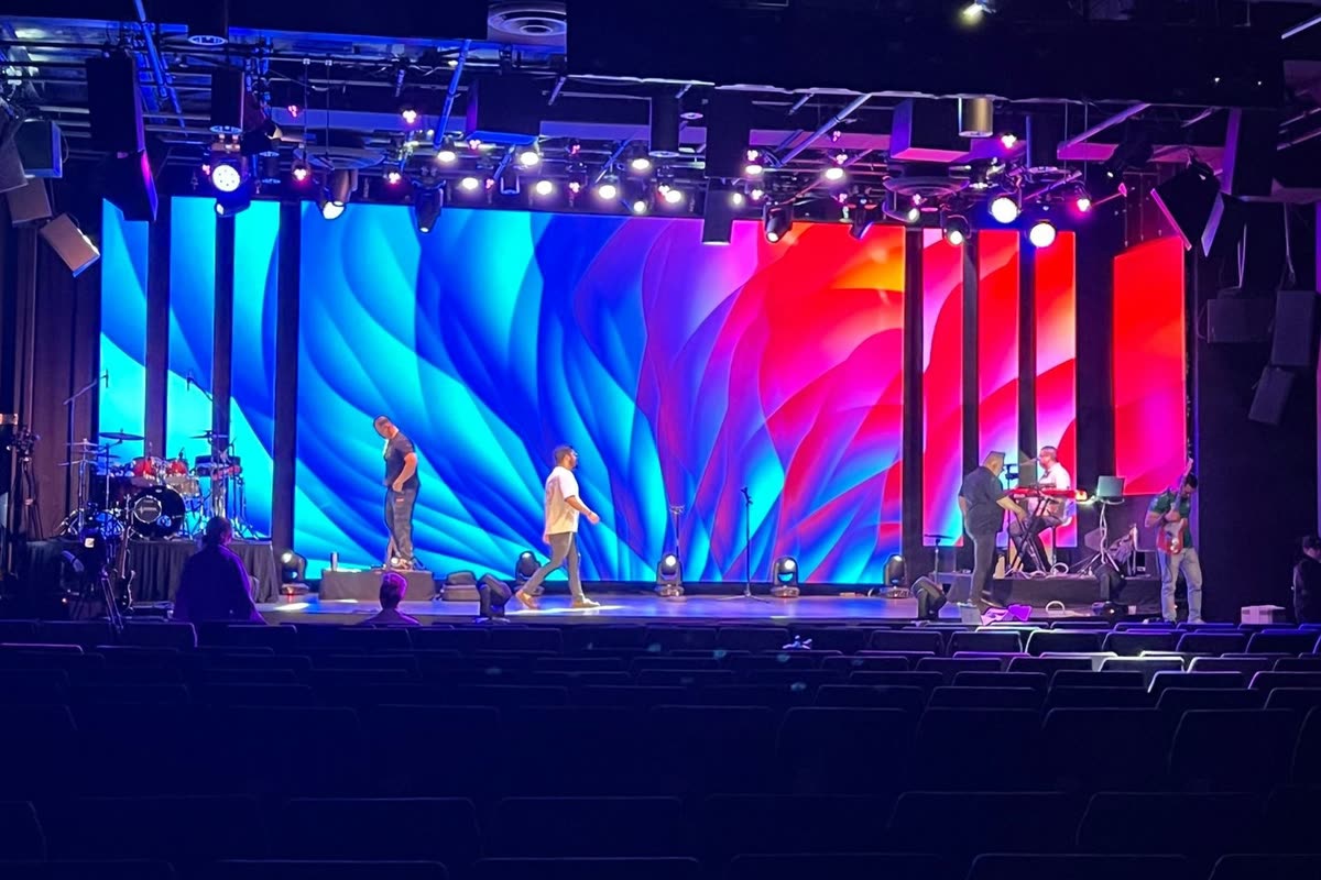 A wide LED wall filling the back of a theater stage with a blue and red abstract graphic, crew walking the empty stage in front of it.