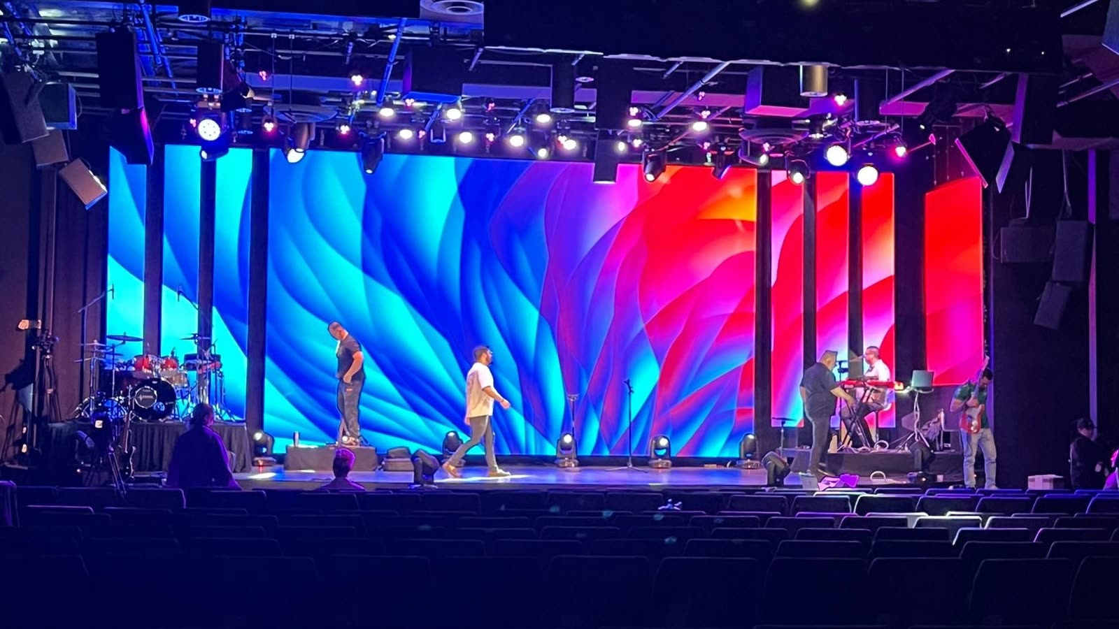 A wide LED video wall filling the back of a theater stage with a blue and red abstract graphic, lit by overhead stage lighting, with empty seating in the foreground.